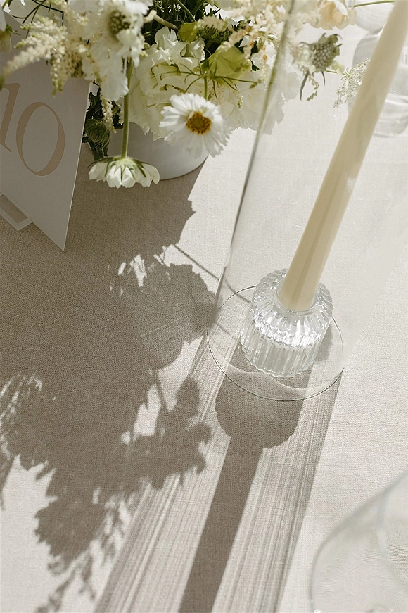 Reception tablescape with wedding table decor, white floral centerpiece, place card, and taper candle in a glass candlestick on linen with sunlit shadows