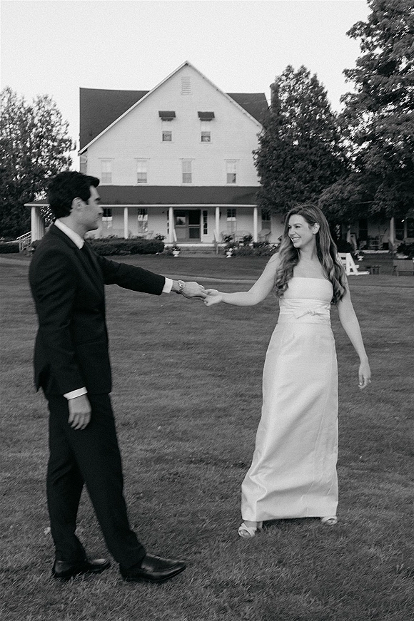 Couple portrait in a black and white wedding portrait style, bride twirling in a strapless dress as groom holds her hand on a farmhouse lawn