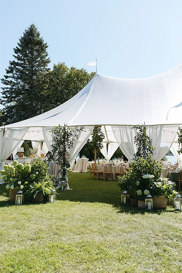 Outdoor wedding tent with sailcloth wedding tent draping, greenery garlands and lanterns, set with long tables on a lakeside lawn