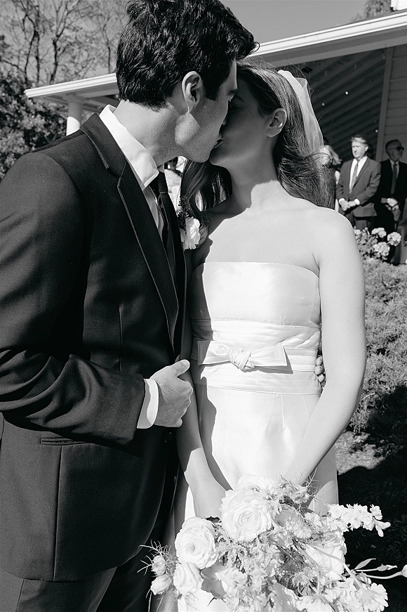 Wedding kiss as the bride in a strapless dress and veil embraces her groom, bouquet in hand, with guests under a pavilion behind