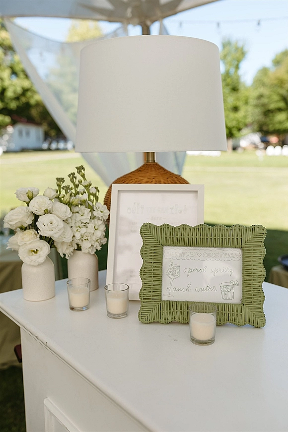 Wedding cocktail signage in a green wicker frame beside a table lamp, white florals and votive candles under a tent with string lights