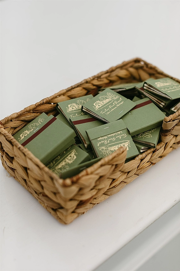 Wedding matchbooks with custom wedding matchbooks gold foil monogram, green covers and burgundy band tucked in a woven basket on a white tabletop