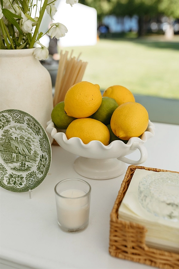 Wedding welcome table with lemons and limes in a white ceramic compote bowl, taper candles and white flowers on an outdoor lawn