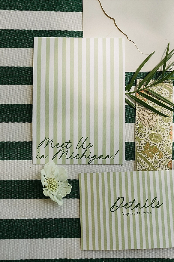 Invitation suite of green striped wedding invitations with envelope and floral insert, styled on green-and-white striped fabric with a white flower