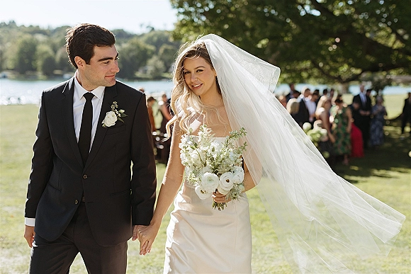 Couple portrait of bride and groom holding hands, her long cathedral veil blowing as they walk on a lakeside lawn with guests behind