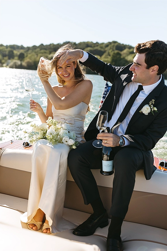 Couple portrait of newlyweds on a boat deck, bride in strapless satin dress holding bouquet and champagne flute, lake behind them
