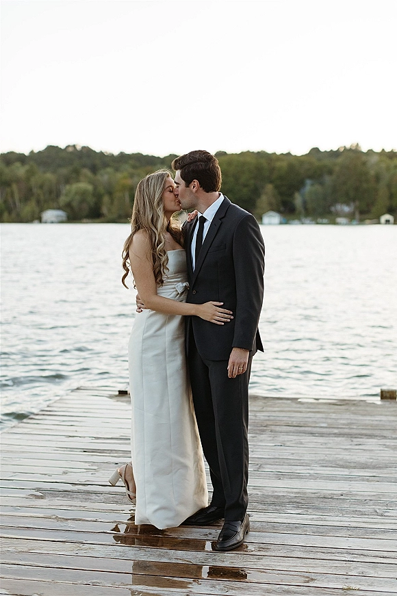 Wedding kiss portrait of bride and groom kissing on a wooden dock by a lake, bride in strapless dress and heels with long wavy hair