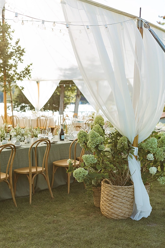 Reception tablescape with outdoor reception tables on green cloth, hydrangea centerpieces, wine glasses and bentwood chairs under a white draped tent with string lights by the water view