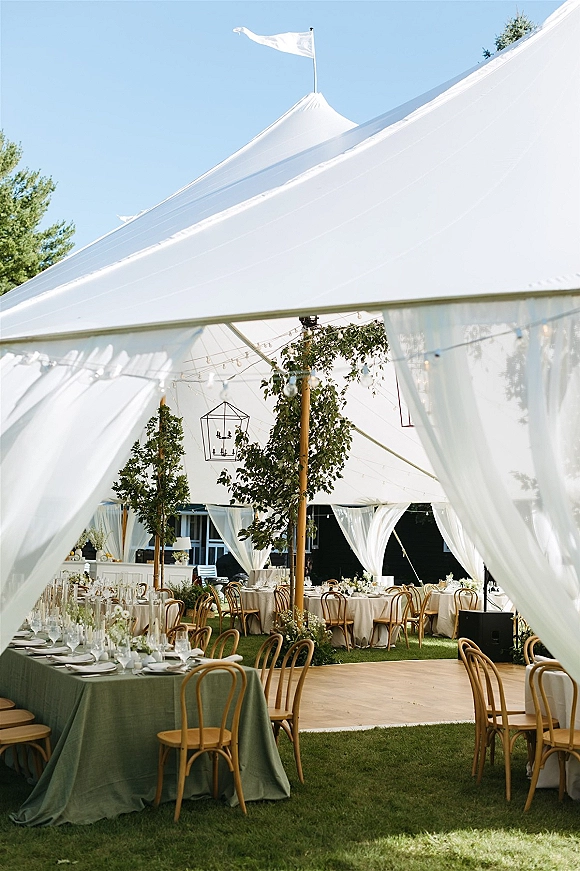 Wedding tent reception under a sailcloth wedding tent with sheer drapery, string lights, lanterns, and round tables on a lawn under blue sky