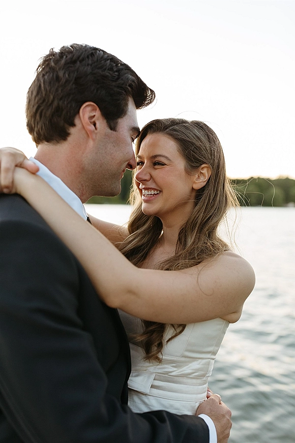 Couple portrait of bride and groom embrace, smiling at golden hour by a lakeside shoreline, her strapless wedding dress beside his suit