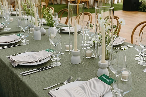 Reception tablescape with green linen tablecloth, white taper candles and bud vases, striped napkins, and handwritten place cards on a long table