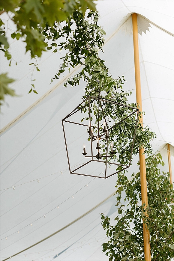 Wedding tent lighting with a geometric chandelier and candles hanging among greenery garlands under a white sailcloth canopy with string lights