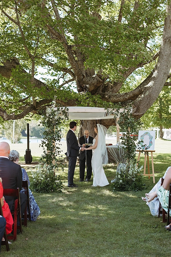 Wedding vows during an outdoor wedding ceremony as bride and groom hold hands under a greenery arch by a lakeside tree in daylight