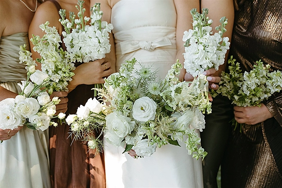 Bridesmaid bouquets of white and green bouquets held by bridesmaids in satin dresses, tied with ribbon, against lush outdoor greenery