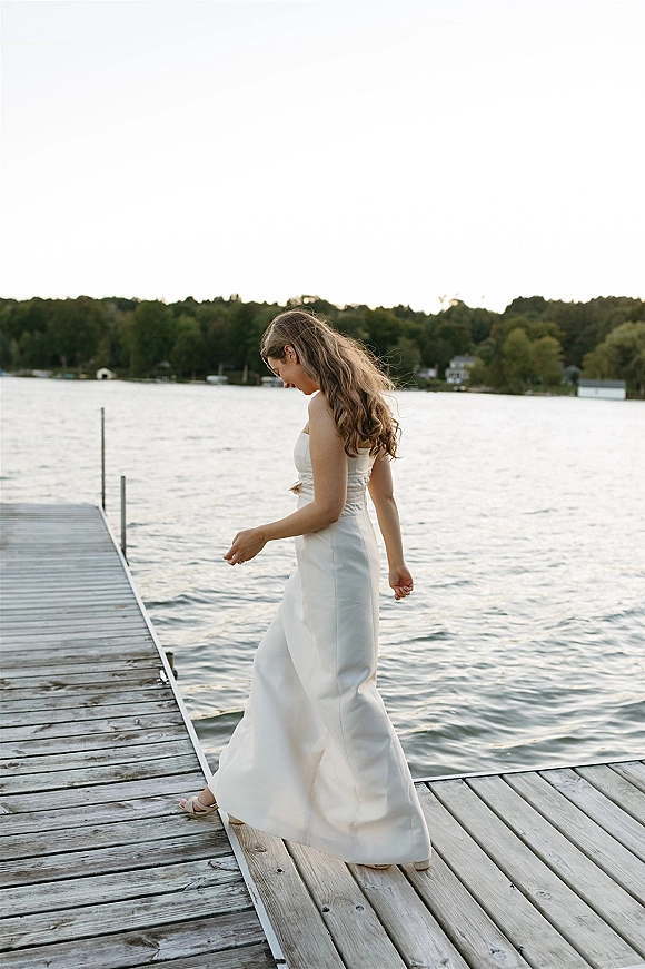 Bridal portrait of a bride walking on dock in a strapless satin wedding dress with thin belt sash and sandals by a calm lake