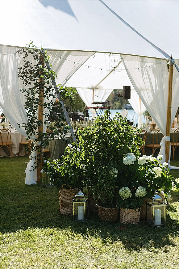 Reception tent decor with a white draped tent, greenery garlands, lanterns and candles, bistro chairs, and string lights by a lakeside lawn