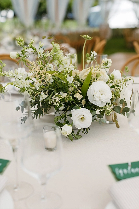 Wedding centerpiece with white floral centerpiece and greenery, votive candles, wine glasses, and place cards on a white tablecloth under a tent