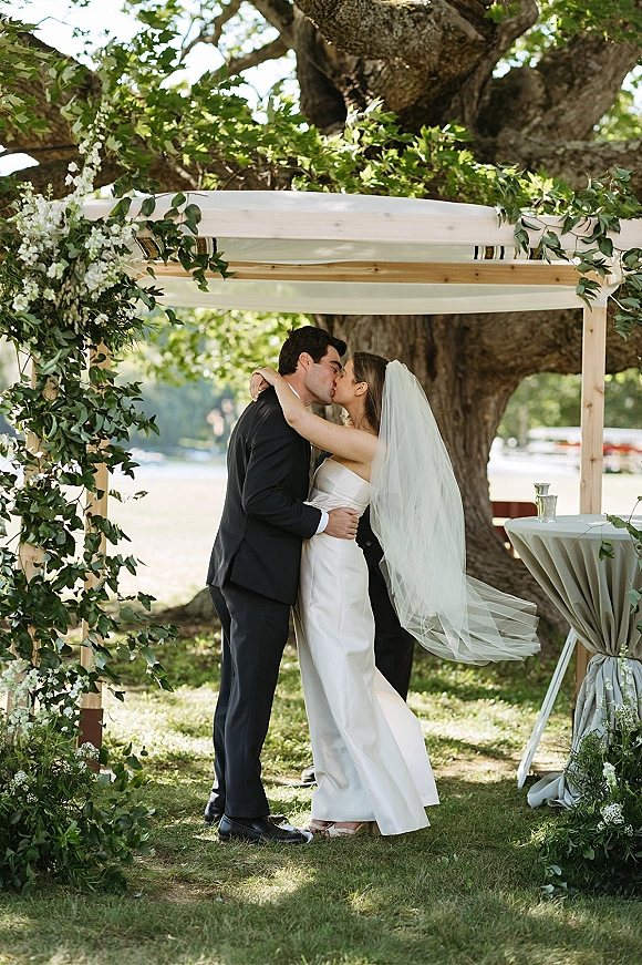 Wedding kiss as bride and groom kiss beneath a greenery wedding arch with a flowing veil on a lakeside lawn under a large tree