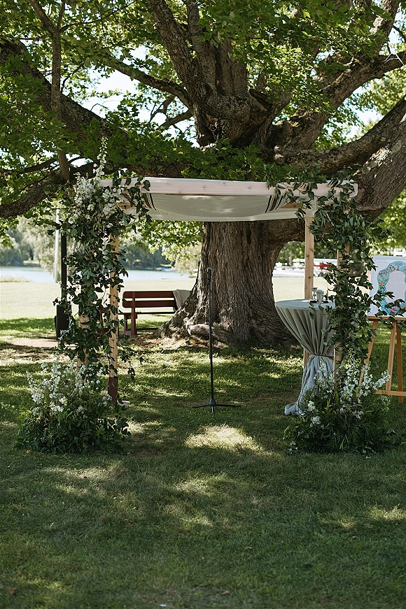 Wedding ceremony arch with a wooden wedding arch, greenery garland and white flowers, fabric drape, beneath a large tree by a lake lawn