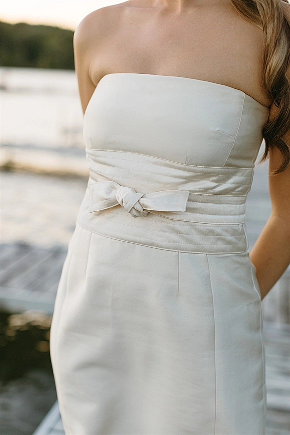 Wedding dress close-up showing a strapless wedding dress in smooth satin with a waist sash and bow belt on a wooden dock by the lake