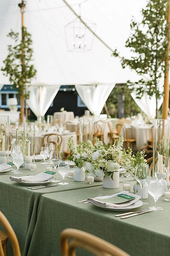 Reception tablescape with green wedding tablecloth, white floral centerpieces and candles, set under a draped outdoor tent with hanging lanterns