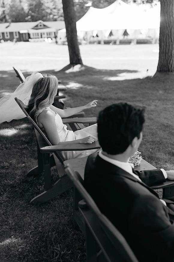 Couple portrait in a black and white wedding photo, bride in veil and groom in tuxedo on Adirondack chairs on a lawn by a tent
