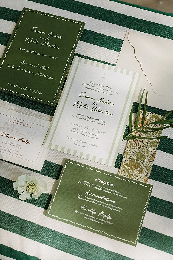 Wedding invitation suite with green wedding invitations, envelopes, ribbon, floral liner, greenery sprig, and white flower on striped fabric