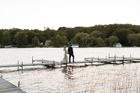Couple portrait of bride and groom on dock holding hands, wedding dress and suit facing a calm lake with boathouse and trees