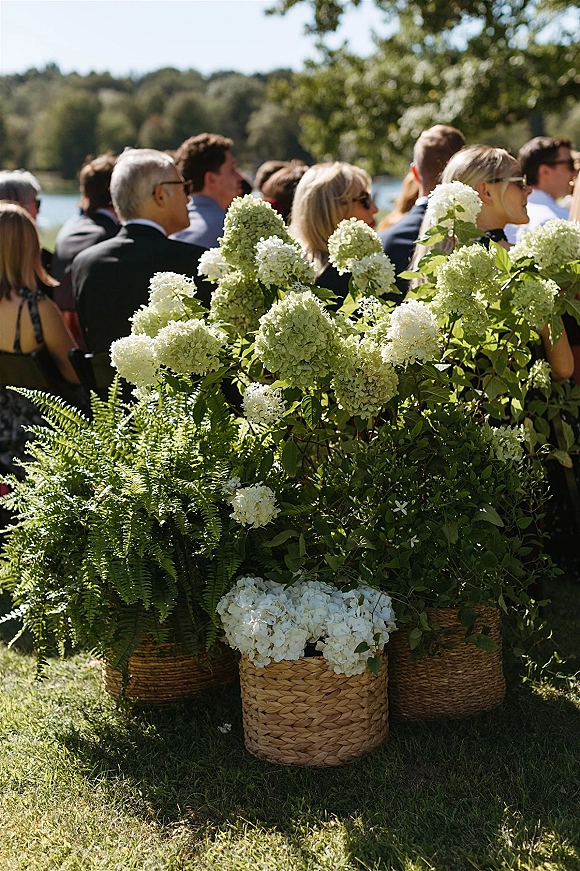 Wedding guests seated at an outdoor wedding, some in suits and sunglasses, by white hydrangeas and fern baskets on a lakeside lawn