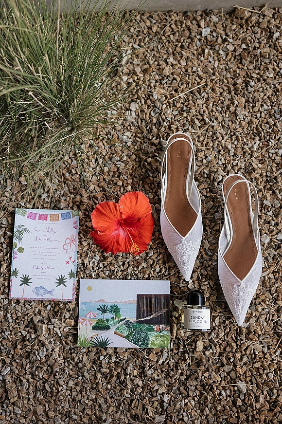 Wedding flat lay featuring a wedding invitation, illustrated postcard, white lace slingback heels, perfume bottle, and red hibiscus on gravel outdoors