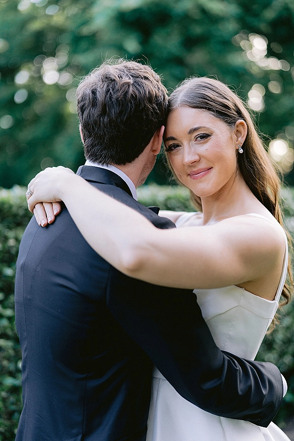 Couple portrait of bride hugging groom, over the shoulder bride portrait looking at camera, pearl drop earrings against garden foliage backdrop