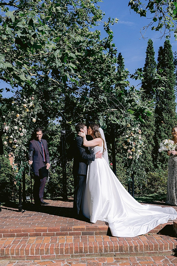 Wedding kiss at an outdoor wedding ceremony under a rose-and-greenery floral arch, bride’s cathedral veil and train flowing on steps