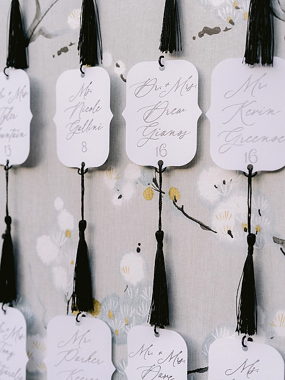 Wedding escort cards with calligraphy on white paper tags, each hung on hooks with black tassels against floral wallpaper backdrop