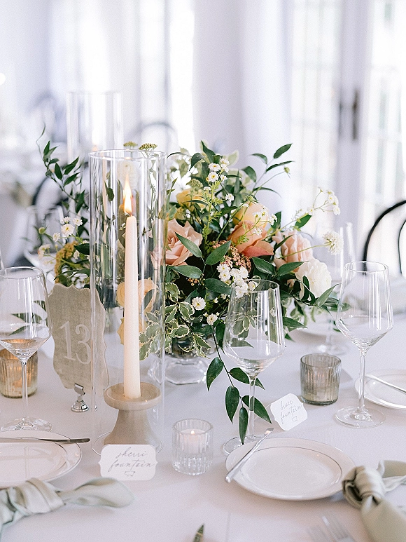 Reception tablescape with wedding table centerpiece of blush roses and greenery, taper candles in glass hurricanes on a white tablecloth in bright window light