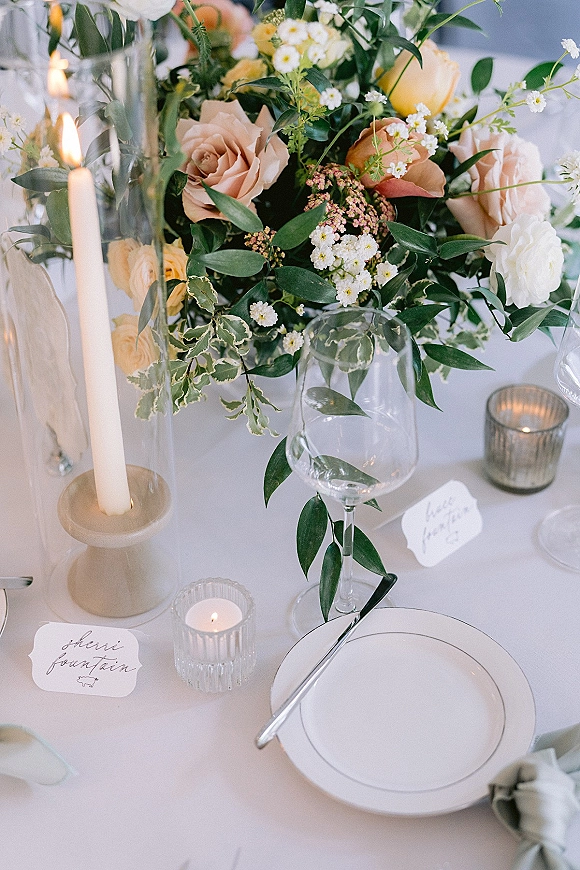 Reception tablescape with wedding table centerpiece of peach roses and greenery, taper and votive candles, and place settings on white linen