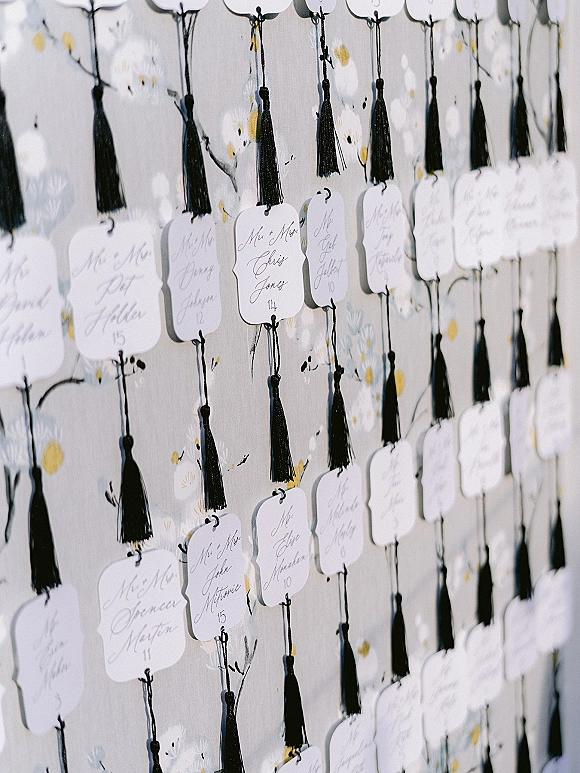 Wedding escort cards with calligraphy names hang on strings with black tassels, displayed against floral wallpaper on a wall display