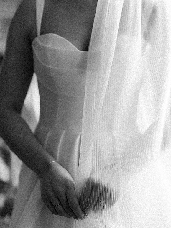 Bridal dress close-up on a strapless wedding dress as the bride’s hands hold the veil, wedding ring visible in soft, blurred interior light
