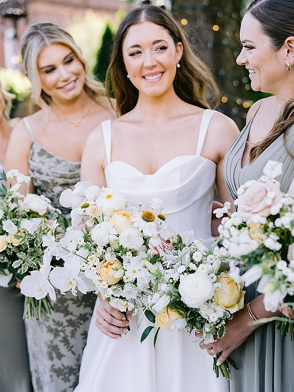 Bride with bridesmaids holding bouquets of white flowers and yellow roses in an outdoor garden with trees and string lights overhead