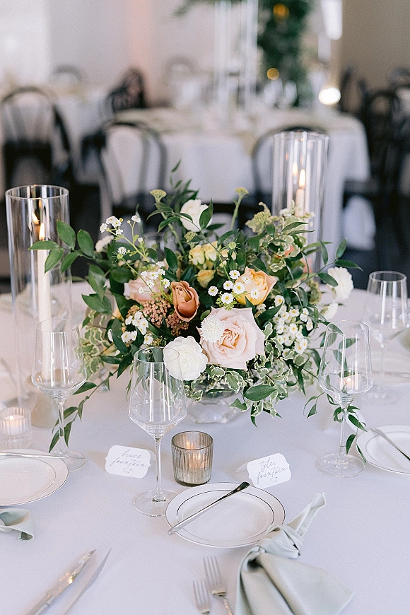 Reception tablescape with wedding table centerpiece of blush roses and white florals, taper candles, glass vases, and place settings on round tables indoors