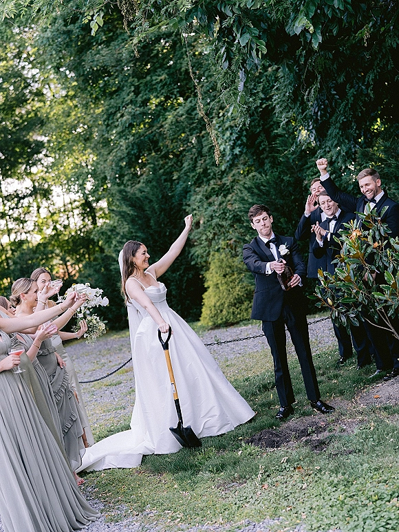 Champagne celebration moment as bride and groom pop a bottle, spraying the cheering wedding party on a tree-lined lawn by a gravel path