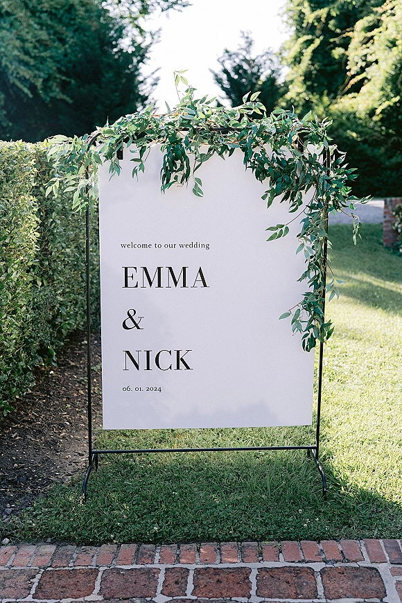 Wedding welcome sign on a black metal stand with greenery garland, set on a brick walkway by hedges and lawn in a garden