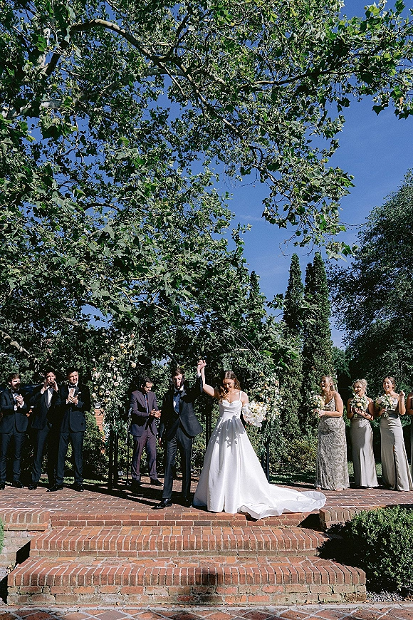 Recessional moment as newlyweds walk the aisle, cheering hand in hand; bride lifts bouquet, long train, garden arch behind