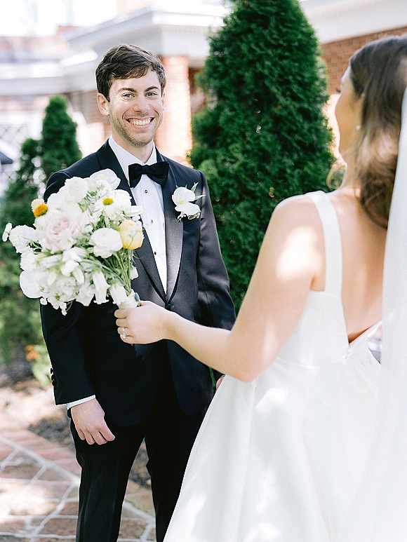 First look moment as groom smiles at bride approaching, bouquet in hand, on a stone walkway by a brick building and shrubs