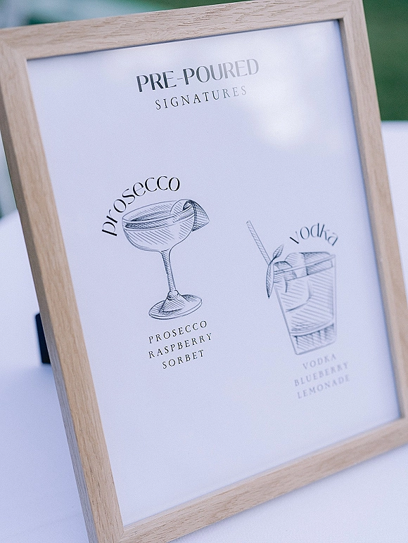 Signature drink sign with wedding signature cocktails in a wood frame, illustrated menu on a white tablecloth with greenery behind
