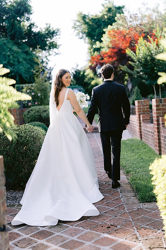 Couple portrait of bride and groom walking away holding hands, bride glancing over shoulder on a brick garden path with veil train