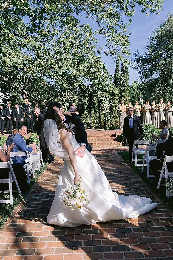 Wedding kiss as the groom dips the bride in a satin gown and veil, bouquet in hand, on a brick aisle at an outdoor garden ceremony