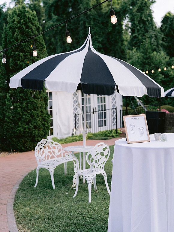 Outdoor cocktail lounge with wedding cocktail hour setup under a black-and-white patio umbrella, string lights, and wrought iron bistro seating on a brick walkway