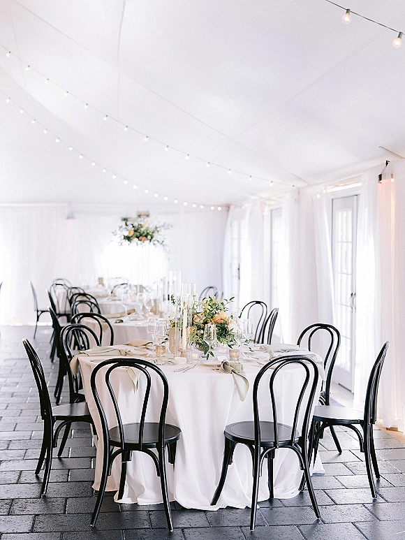 Reception tablescape with round tables, white tablecloths, black bentwood chairs, greenery centerpieces, and taper candle accents under string lights in a white tent