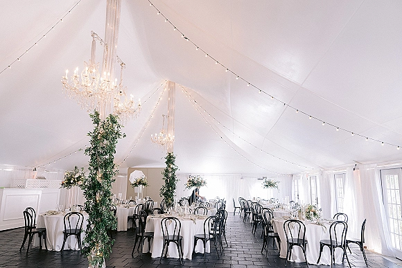 Reception decor in a wedding tent reception with string lights, crystal chandeliers, greenery garlands, and candlelit round tables under draping