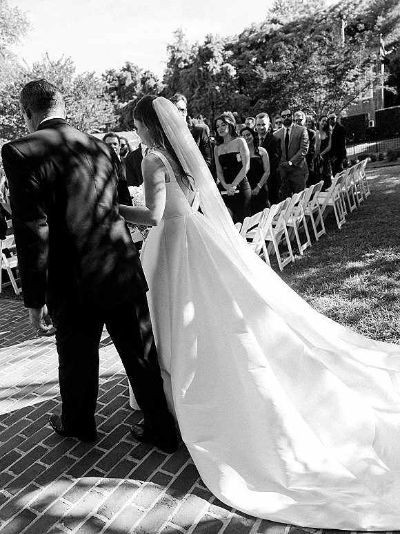 Wedding ceremony moment as bride with a long veil and train walks up a brick walkway aisle past standing guests on a garden lawn